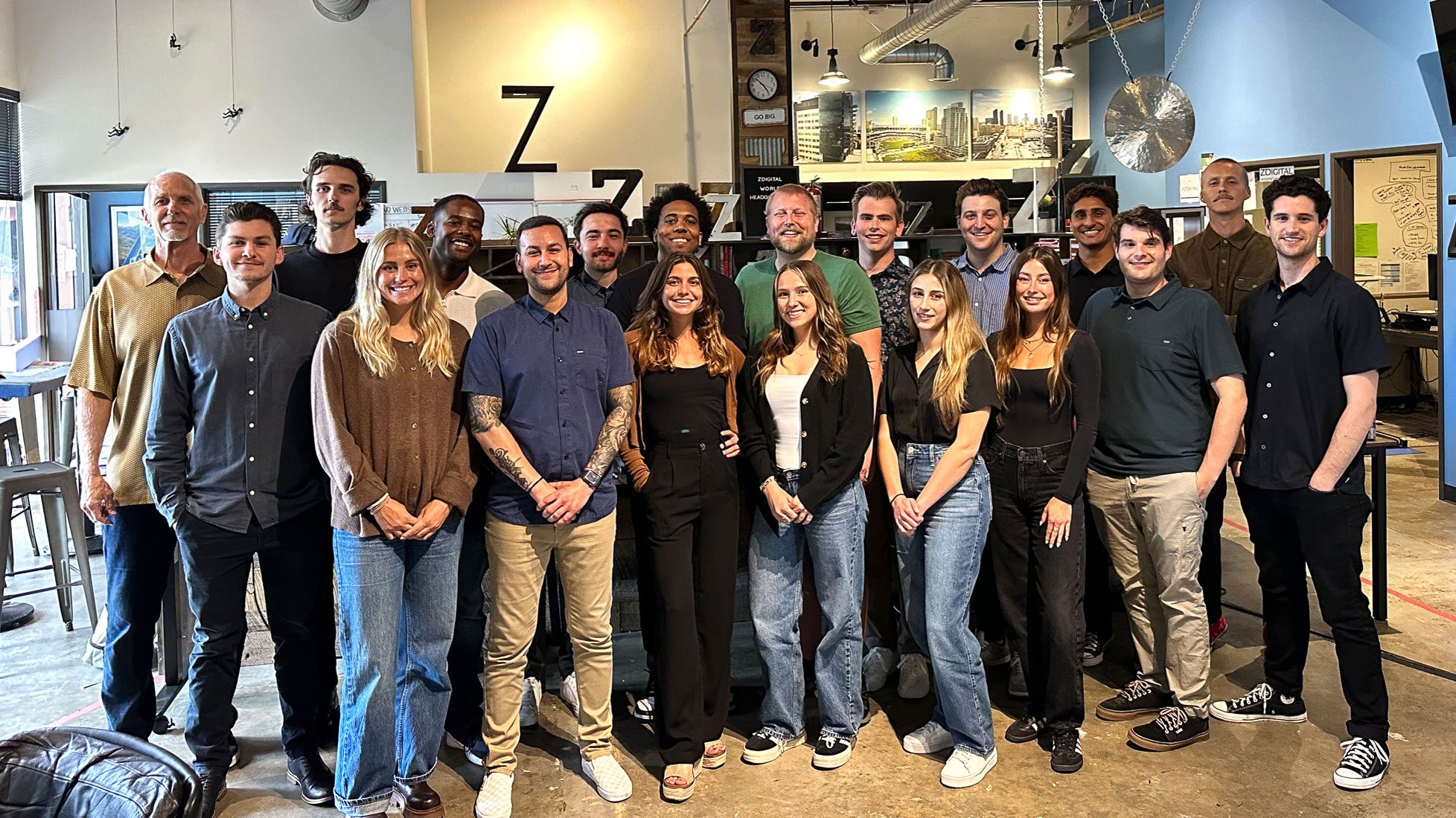 A group of 20 people stand indoors, facing the camera and smiling. They are posed together in a casual setting with various decor elements and furniture in the background.